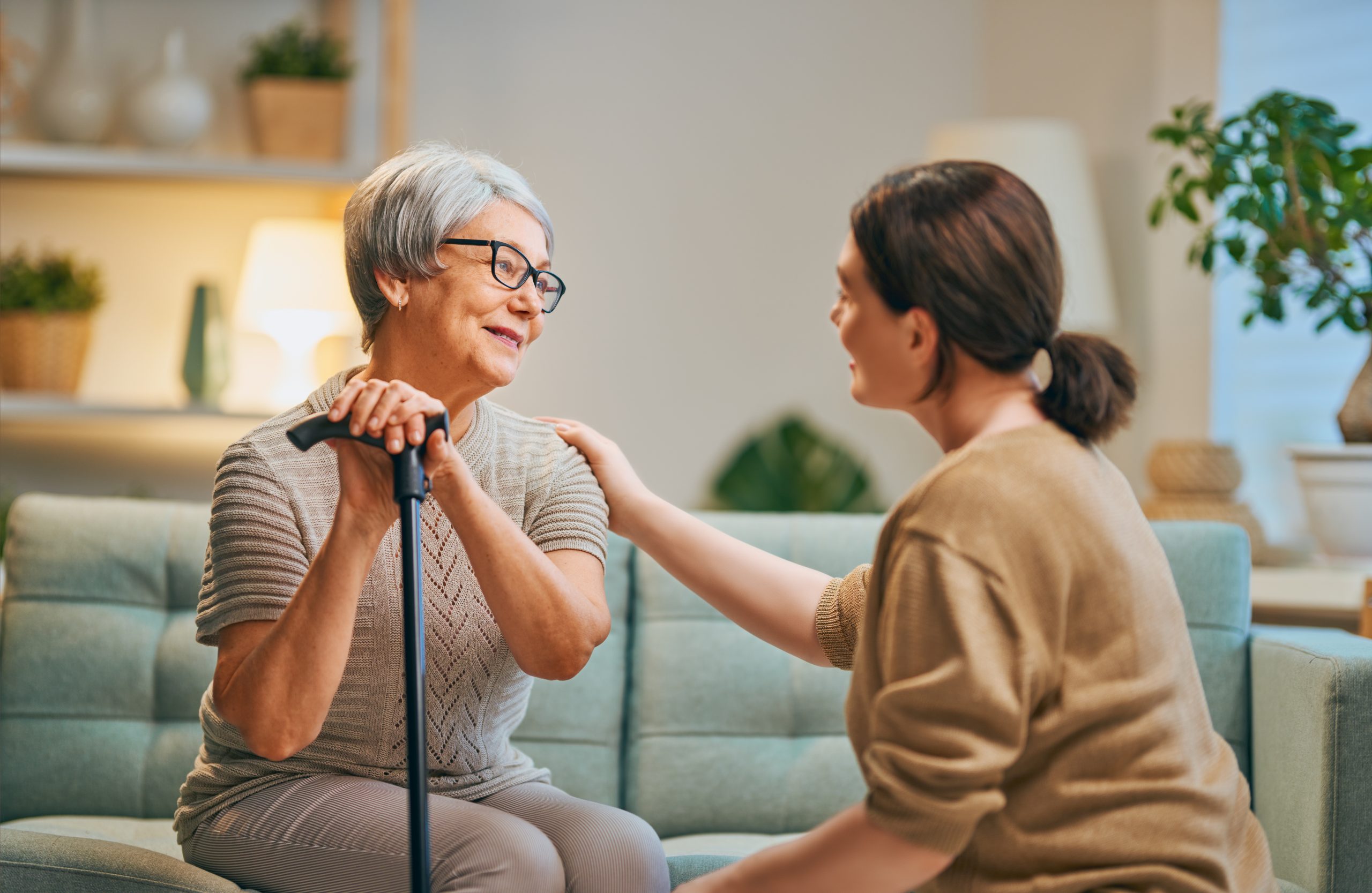 A elderly woman sits on the couch in her home as a young woman embraces her from behind