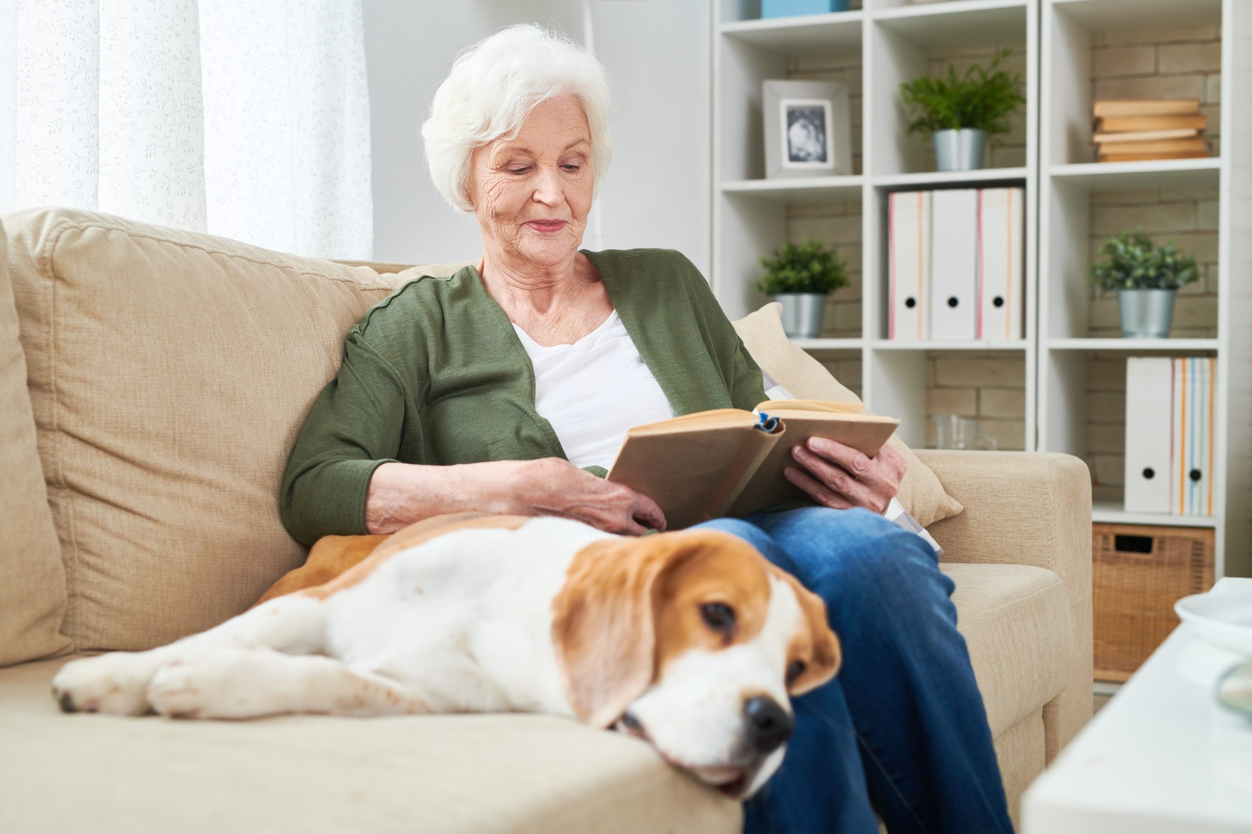 a senior woman reads a book on her couch next to her dog enjoying the many freedoms of in-home care