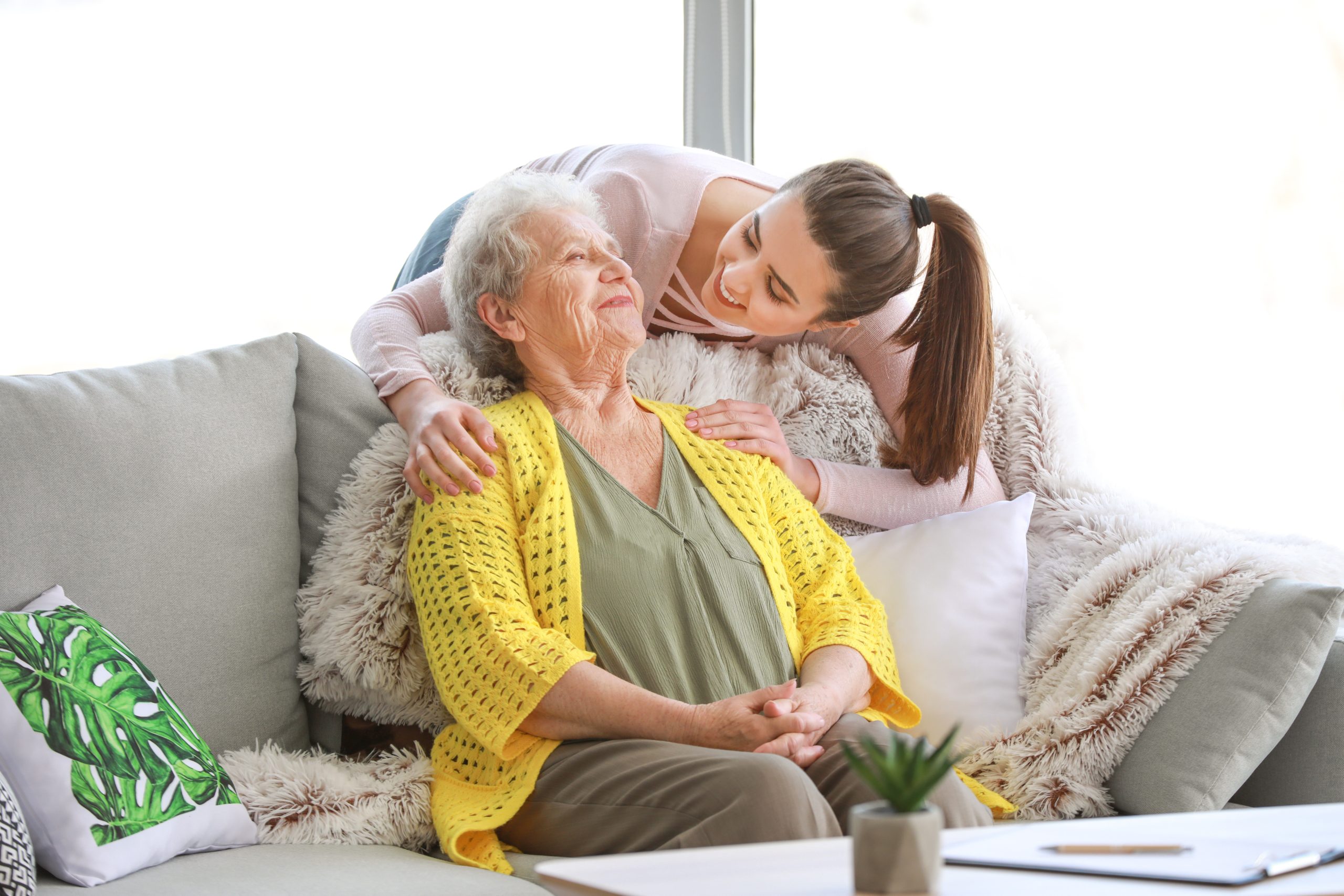 a senior woman and her granddaughter smile on the couch as they talk about the many benefits of in-home care