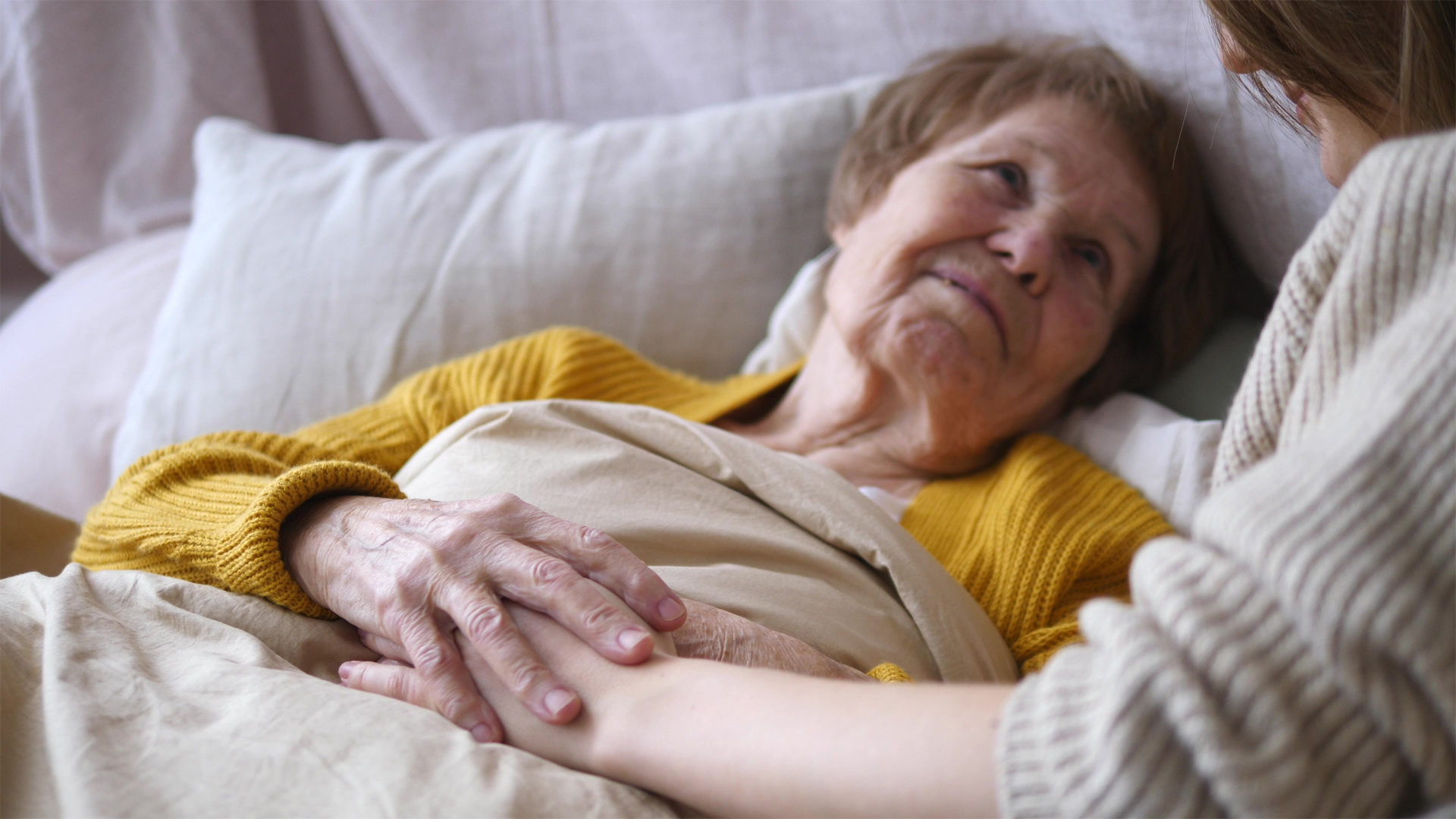 An elderly woman lying in bed touching the hand of the overnight care provider who looks after her during the night.